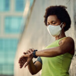African American female runner with face mask using fitness trac