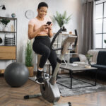 Young african female athlete wearing sportswear and using exercise bike using smartphone.